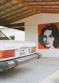 Close-up of the rear of a classic white vintage Mercedes-Benz 380SL with a Texas license plate, parked in a carport next to a large red pop art mural of Elizabeth Taylor. Fine art photography print by Bethany Young.