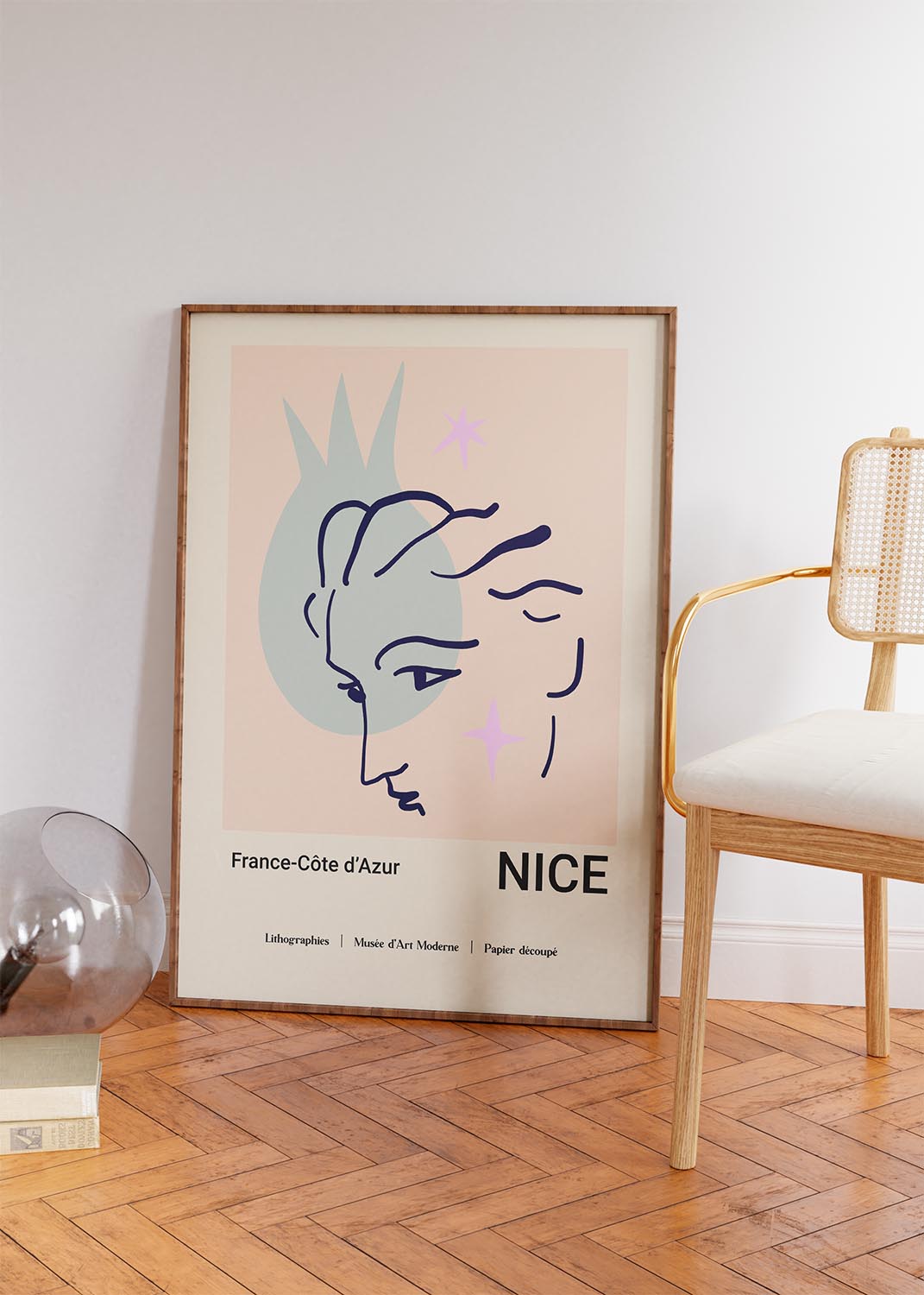 Interior design featuring a framed poster with a Matisse-inspired 'Papiers Découpés' design, depicting a stylized blue profile and pink stars against a peach backdrop, with 'NICE France-Côte d'Azur' text, elegantly placed next to a wooden chair and glass table lamp