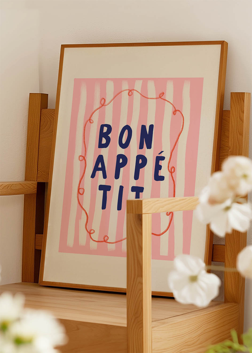 A poster with a light pink background featuring the phrase "Bon Appétit" in the center in large, dark blue letters. The background has a subtle white brushstroke pattern, and a whimsical orange line mimics the outline of an ornate frame, looping around the phrase playfully.