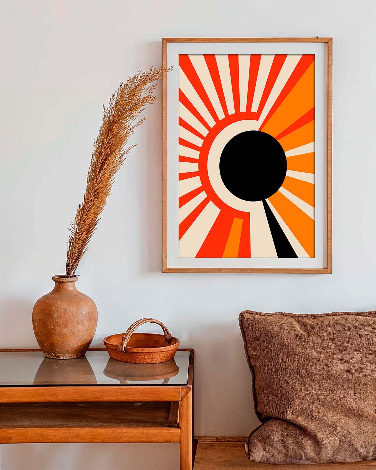 A minimalist art poster displaying a stylized mid-century sunrise with a large black circle at the center, surrounded by radiating orange and yellow rays against a crisp white background, framed and hung on a wall above a wooden console table.